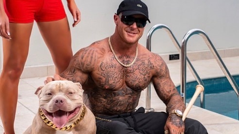 A heavily tattooed man holding a baseball bat sits next to a dog and a pool.