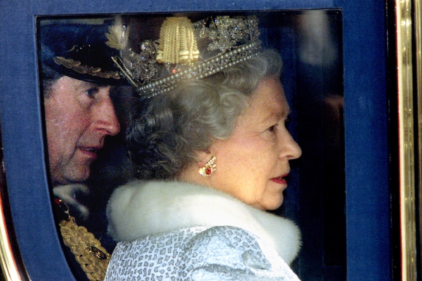 Queen Elizabeth in a fur coat and crown sits in a carriage next to her son Charles 