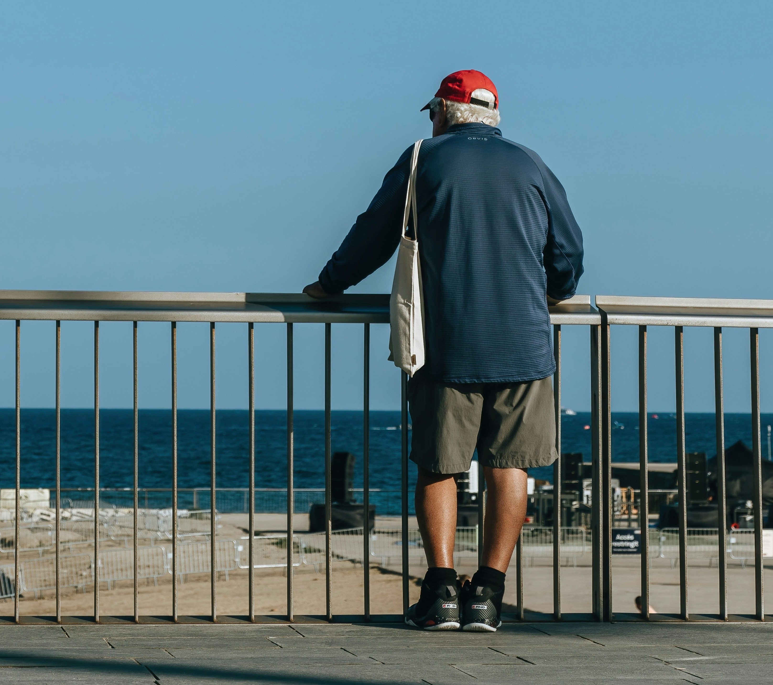 an Older man seen from the back stands at a railing and looks out to sea. 