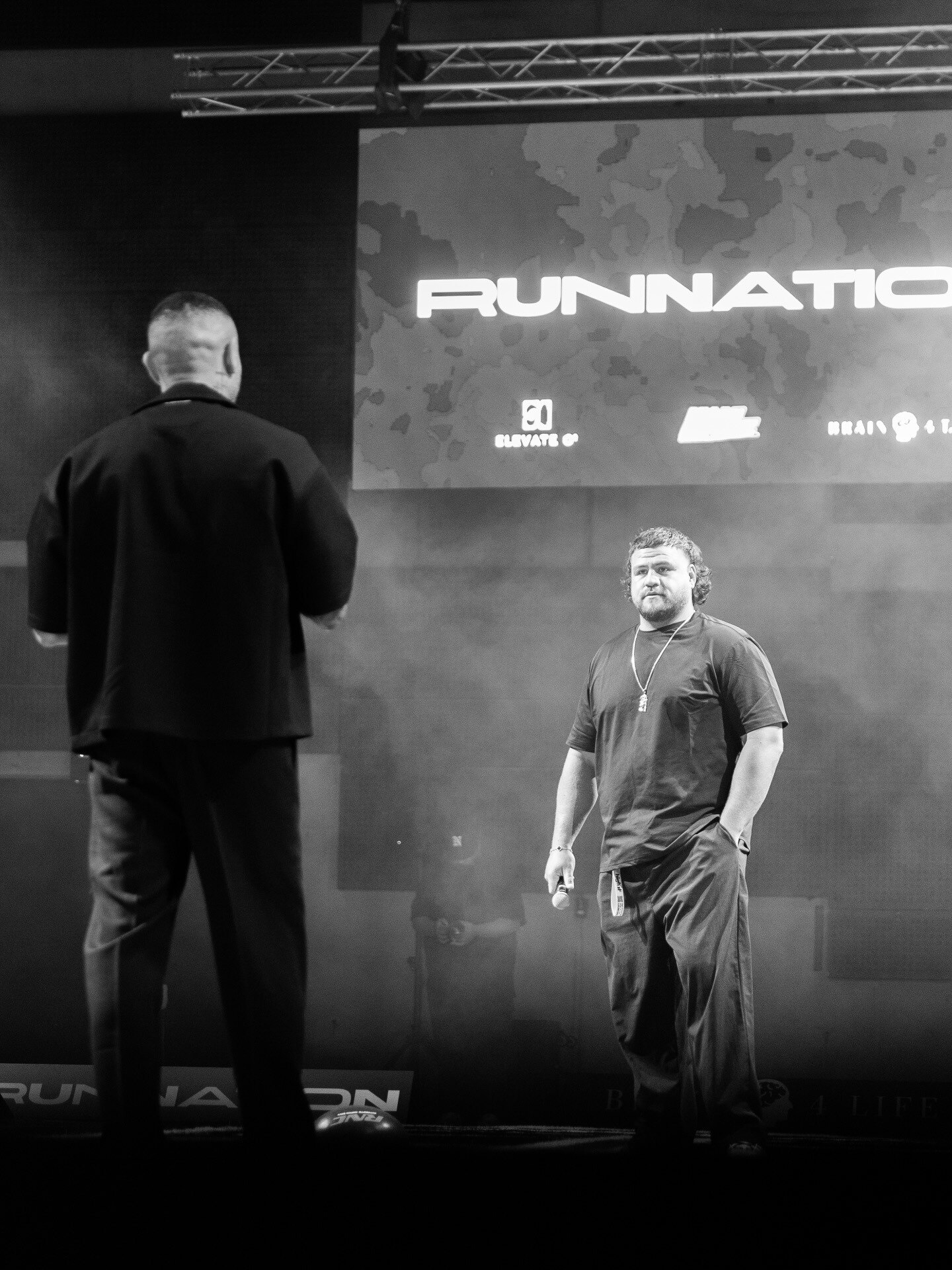 A black and white photo of a man facing another man wearing black and has his hands in his pocket under the sign RunNation