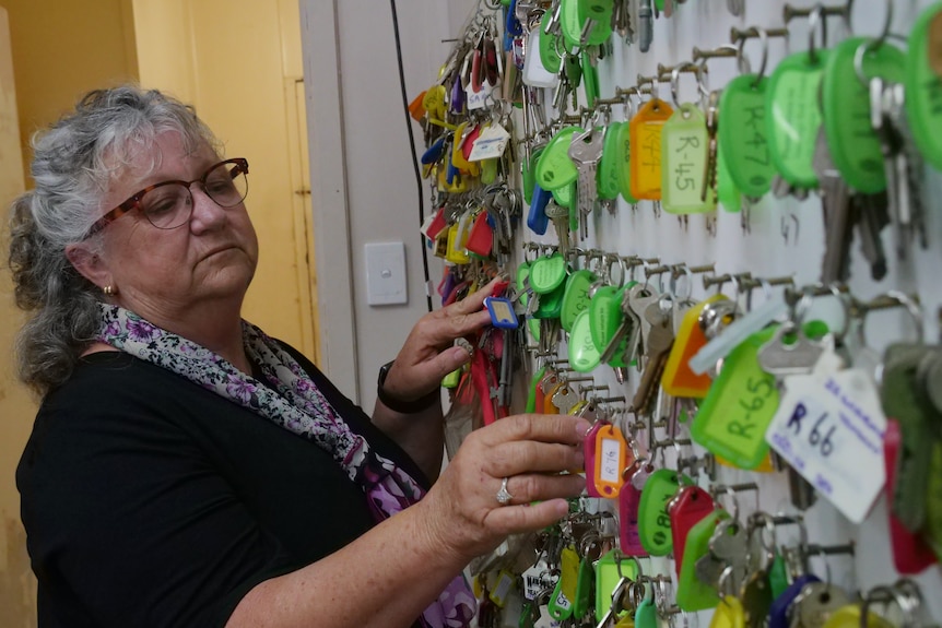 A bespectacled, grey-haired woman puts a set of keys on a wall-mounted rack.
