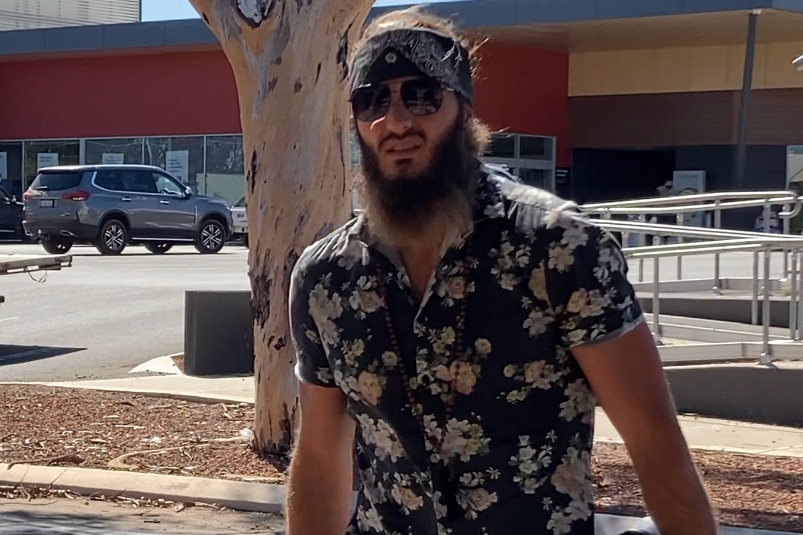 A man with a beard in a floral shirt, sunglasses and with a dark bandana tied around his head stands in a supermarket carpark.