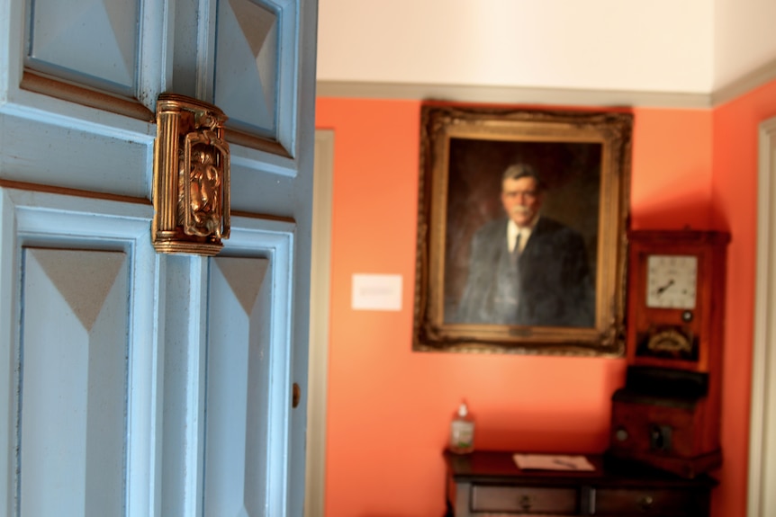 An open blue door with a brass knocker with a portrait out of focus in the background.