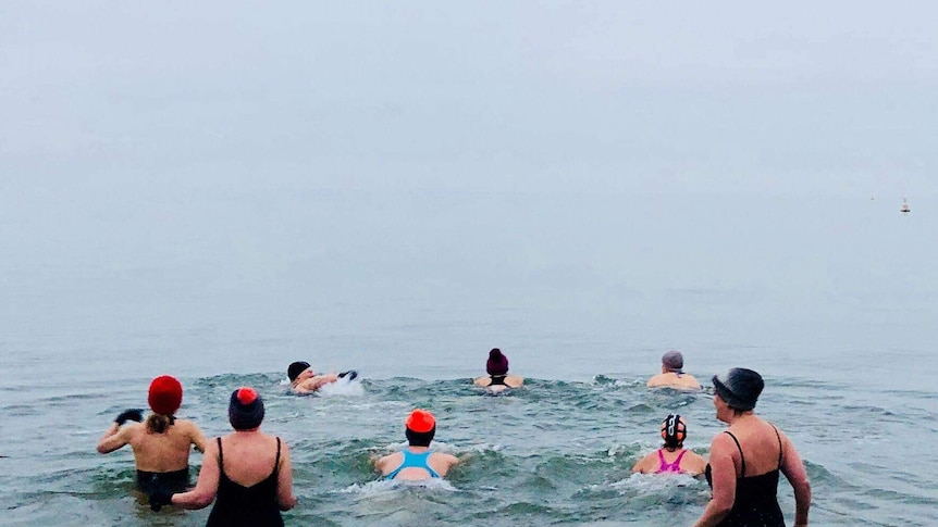 A shot from the beach looking at a group of swimmers wading into cold ocean waters on a grey winter's day