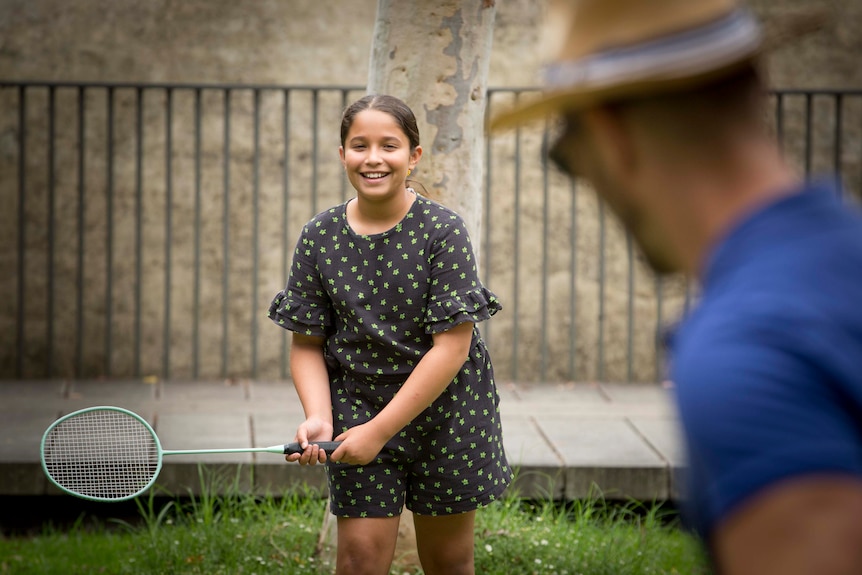 A ten-year-old girl is seen over her father's shoulder holding a badminton racquet. 