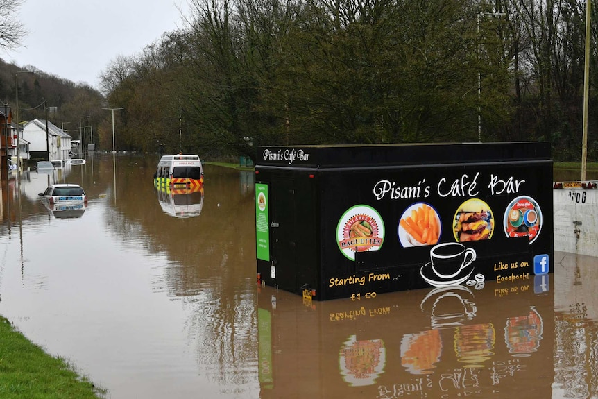 A colourfully painted food van is half-submerged, near two other vehicles and houses, in a flooded street in a Welsh village.