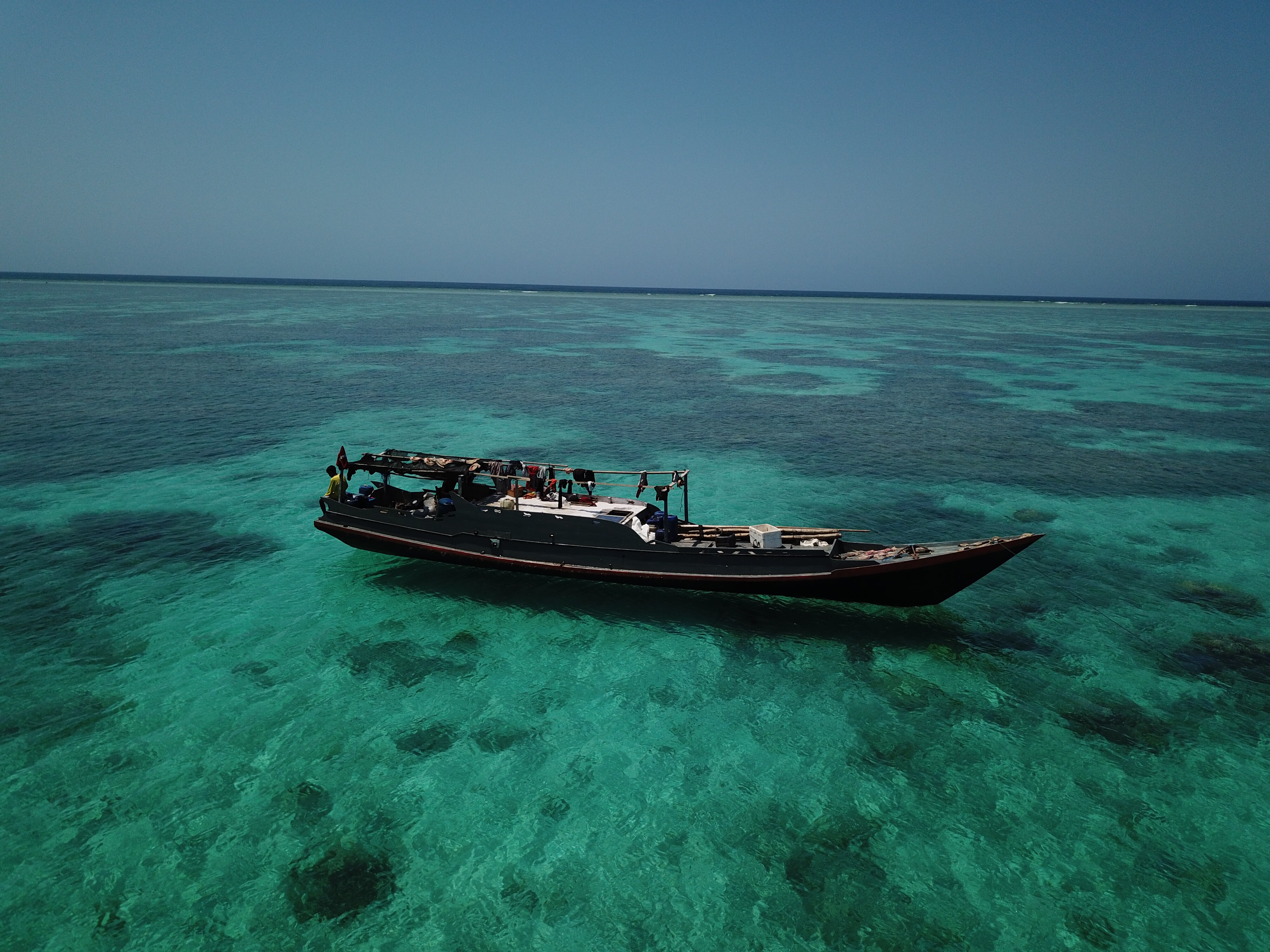 A man sitting on the edge of a long wooden fishing boat, floating on the water on top of a reef.