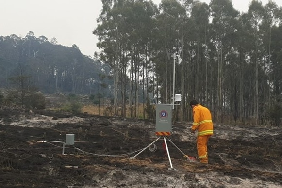 A man sets up monitoring equipment on a hill near some trees.