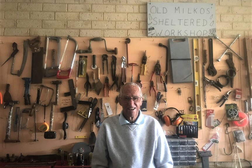 An ninety year old man standing smiling in front of his workshop. The sign reads Old Milkos Shop