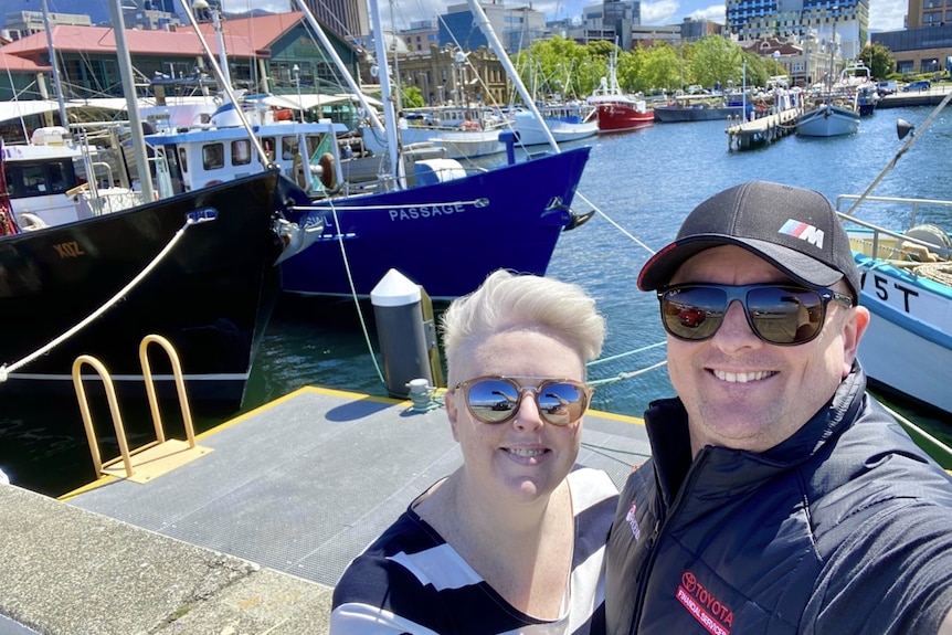 A woman with blonde hair and sunglasses poses for a photo in a boat harbour with a man in a cap