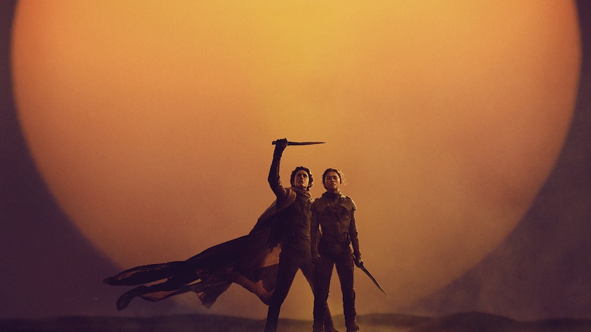 Two people standing in a desert space with a yellow dusty background. One person has their right arm up holding a sword. 