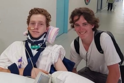 A boy with a neck brace and a sling next to another boy with a backpack on.