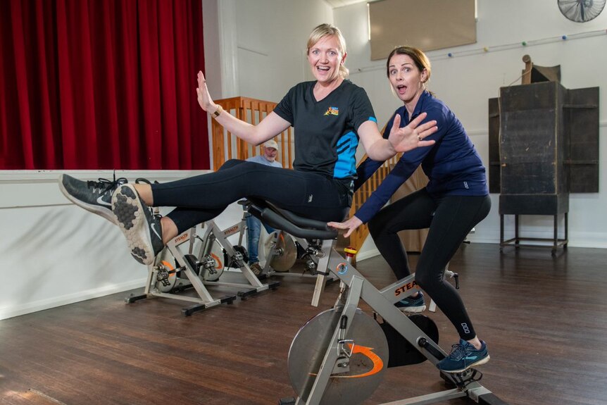 Two women wearing activewear. One sitting on an exercise bike seat and the other on handlebars with arms outstretched laughing.