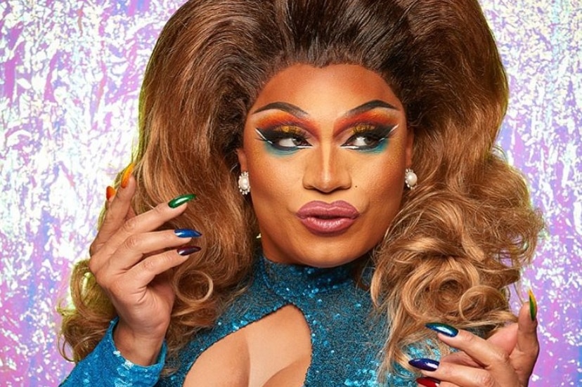 Brita wears a big brown wig with heavy red and green eye makeup, rainbow finger nail extensions and a sparkly turquoise jumpsuit