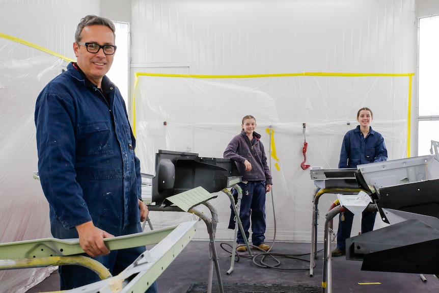 Male teacher in blue overalls with two female students in an auto workshop.