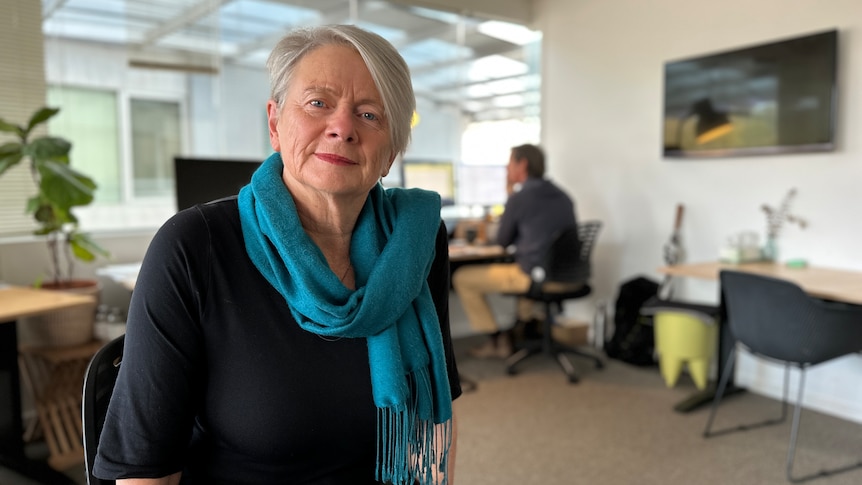 A lady with a blue scarf sitting in an office space with someone working in the background.