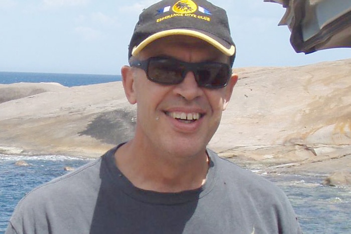 A man in a dark t-shirt and hat stands posing for a photo and smiling standing on rocks in front of the ocean.