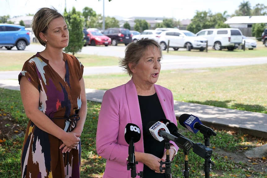 A woman in a brown dress and a woman in a black top and pink jacket standing outside in front of microphones.