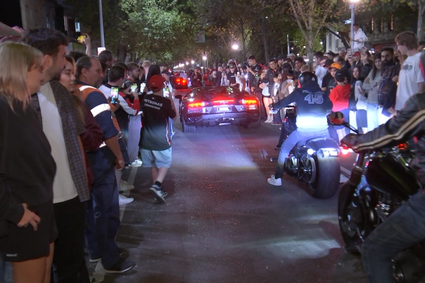 A black Ferrari is followed by two motorbikes down a dark street lined with people.