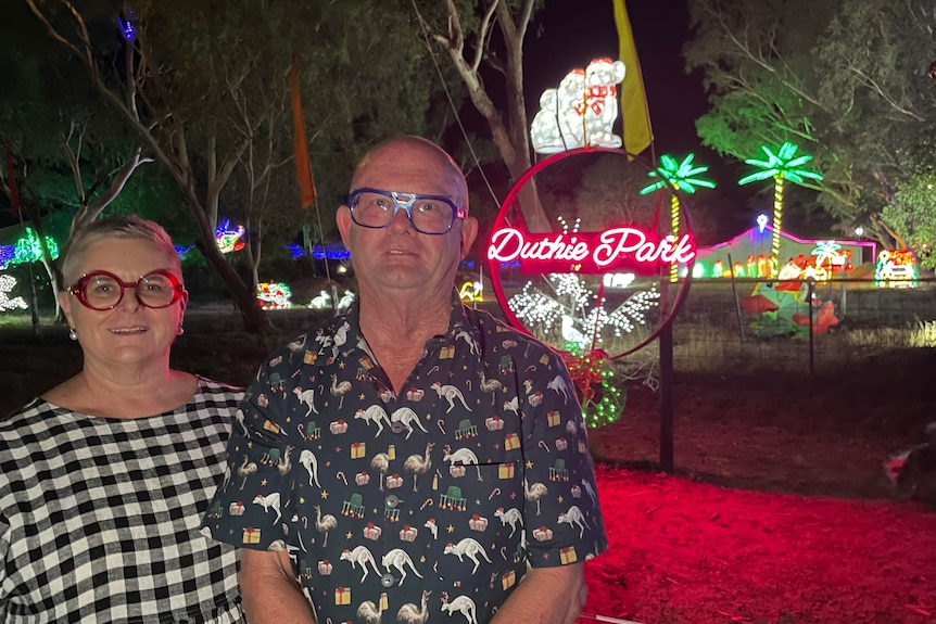 A man and a woman stand in front of a bright Christmas lights display including LED palm trees.