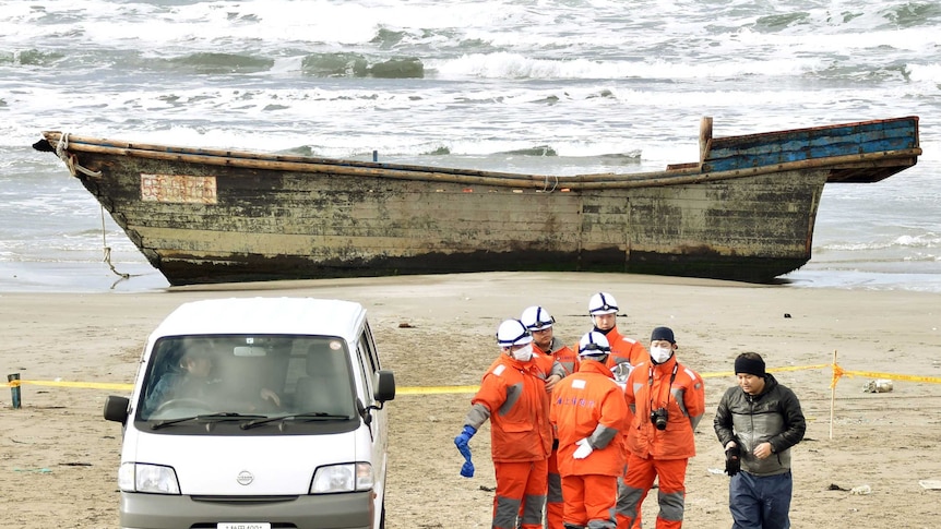 Men in orange jackets stand in front of a wooden boat on a beach.