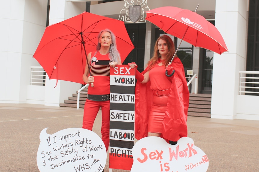 Two women stand holding red umbrellas and placards. 