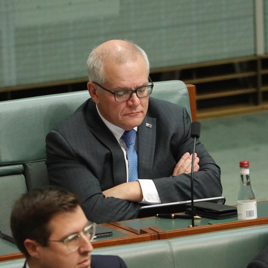 Morrison sits on the green bench in the back row of parliament with his arms crossed