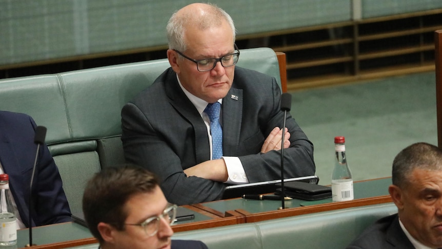 Morrison sits on the green bench in the back row of parliament with his arms crossed