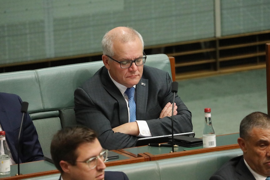 Morrison sits on the green bench in the back row of parliament with his arms crossed