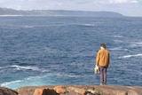 A young man stands on a rock near the edge of the ocean.