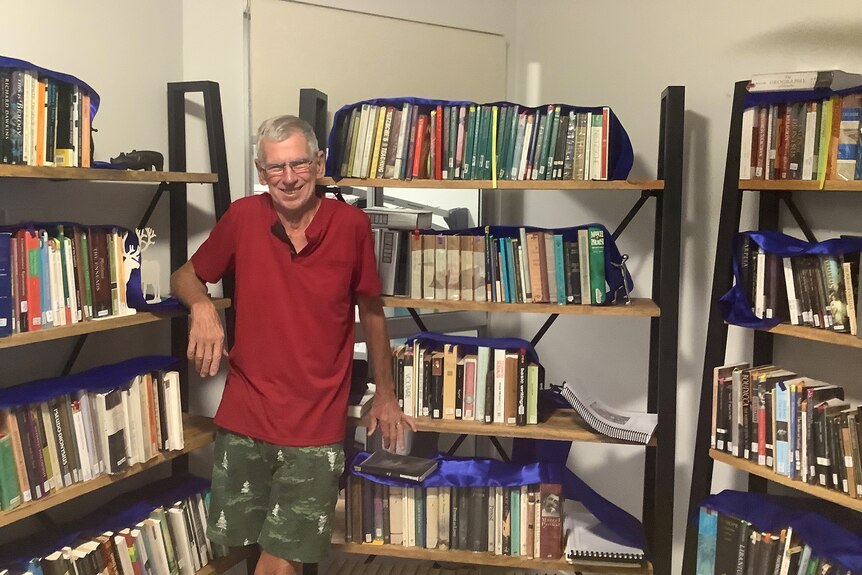 A man with short grey hair leans against two bookshelves while smiling at the camera.