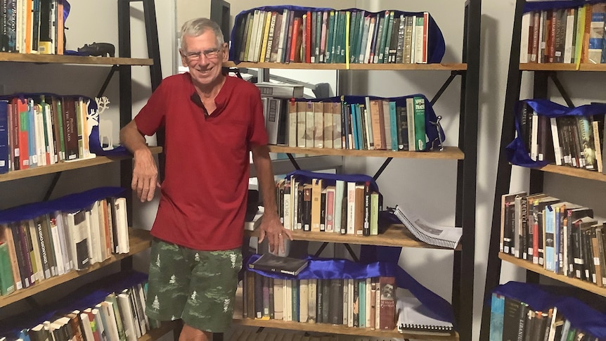 A man with short grey hair leans against two bookshelves while smiling at the camera.
