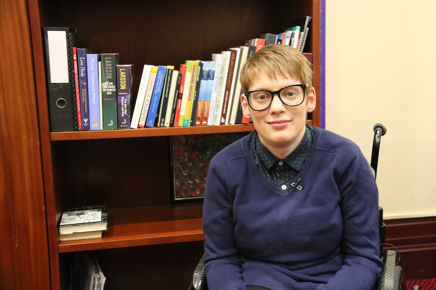 A young woman with short blonde hair and glasses sits in a wheelchair in front of a book shelf in an office.