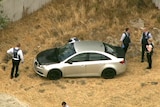 An aerial shot of a silver car with a black bonnet and boot surrounded by police officers parked in a vacant lot.