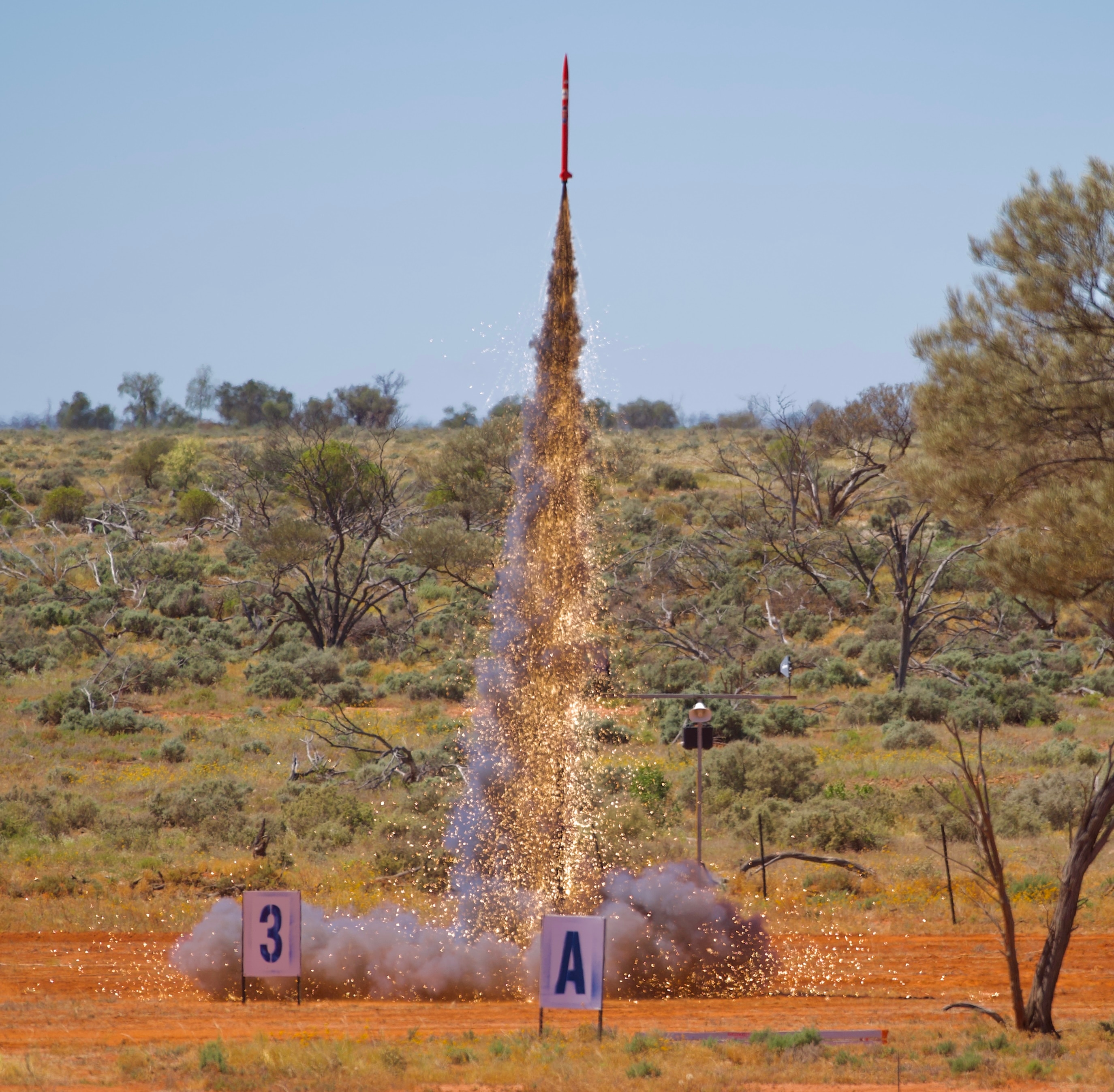 A small, slim rocket leaves a trail of smoke and sparks at is launches into the air in an outback area.