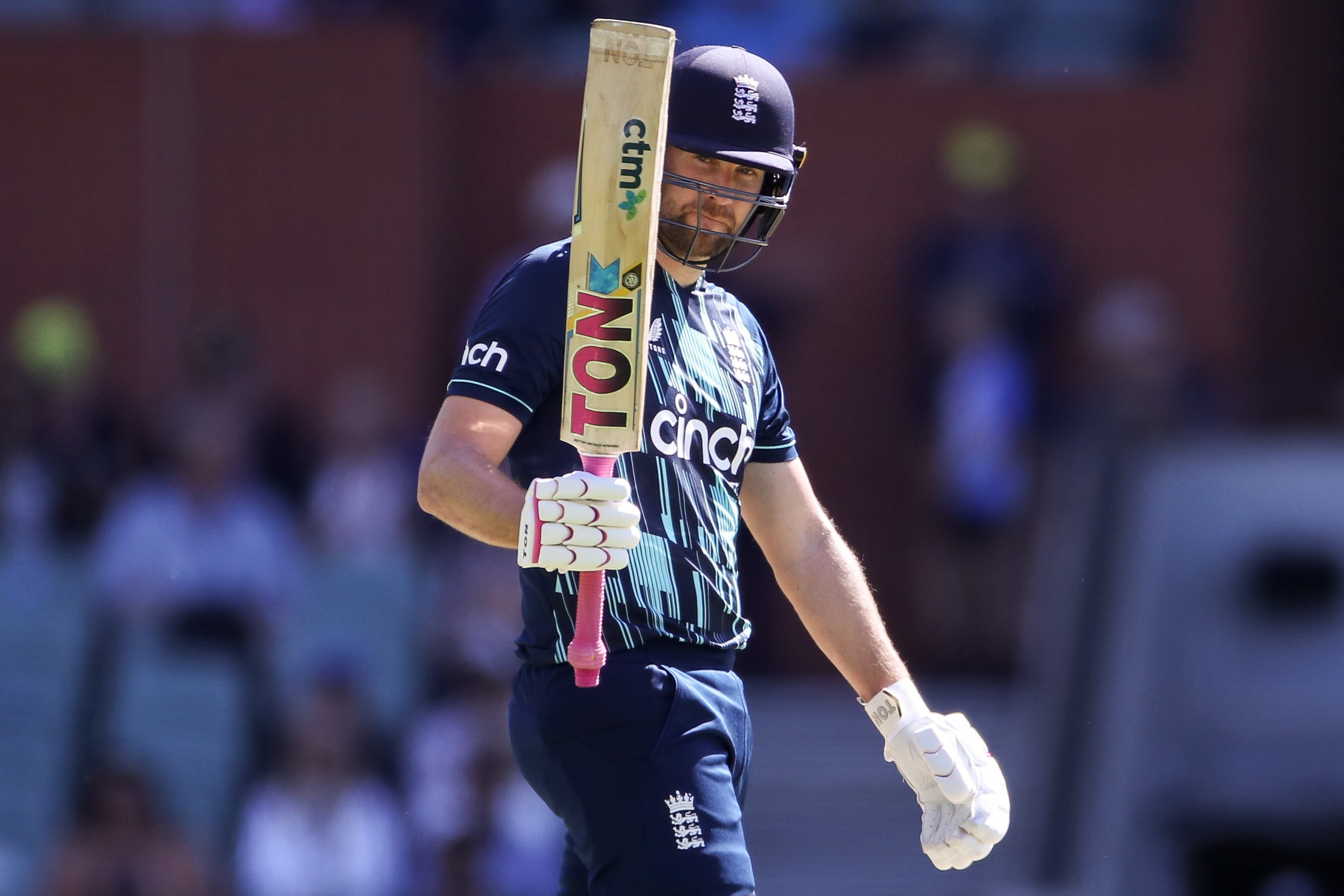 An England male batter holds his bat up with his right hand after scoring a century.