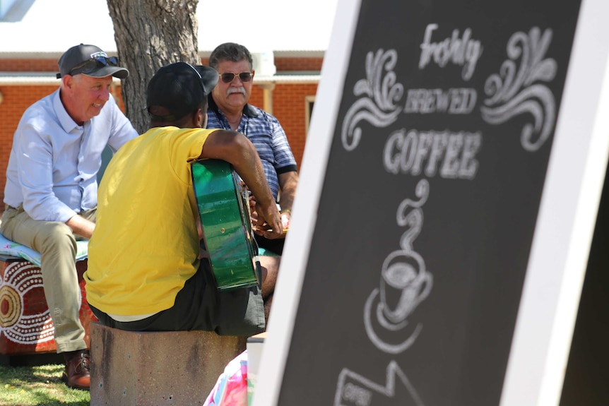 A detainee plays guitar while sitting next to Corrections Minister Fran Logan, with a cafe sign sitting in the foreground.