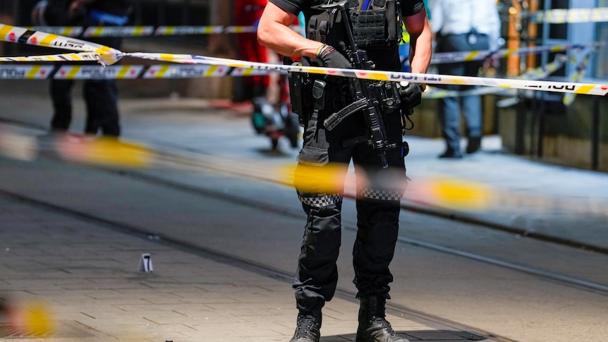 a security guard is seen on a street behind police tape with a rifle in his hand