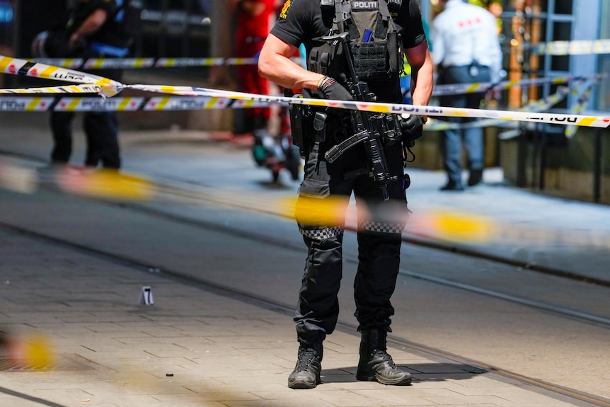 a security guard is seen on a street behind police tape with a rifle in his hand