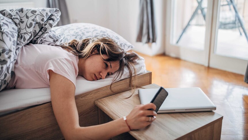 A woman looks at her phone while lying in bed, looking exhausted