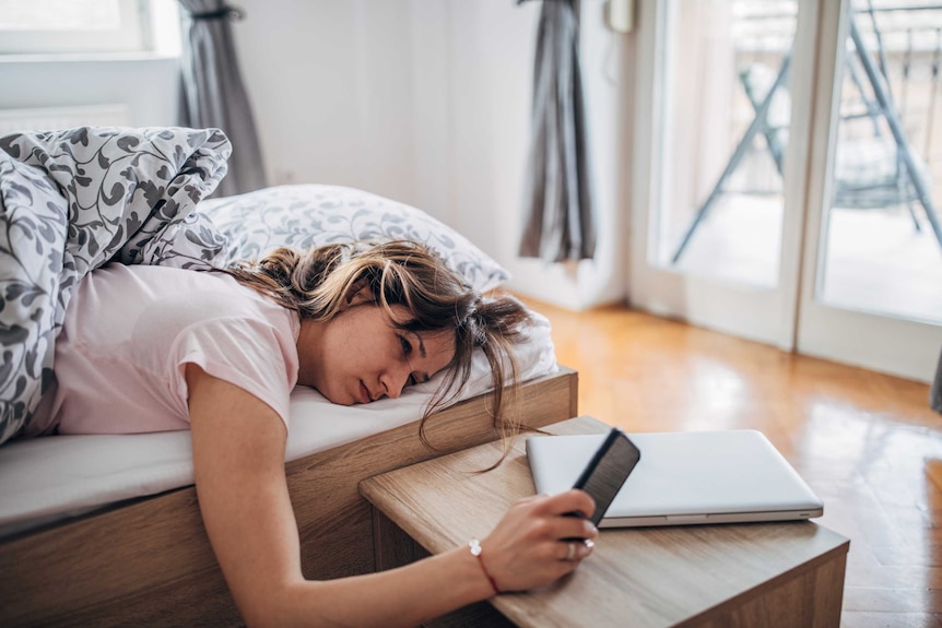 A woman looks at her phone while lying in bed, looking exhausted