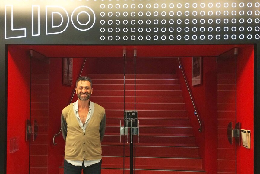 A bearded man, smiling, stands in front of a set of red steps leading into a cinema.