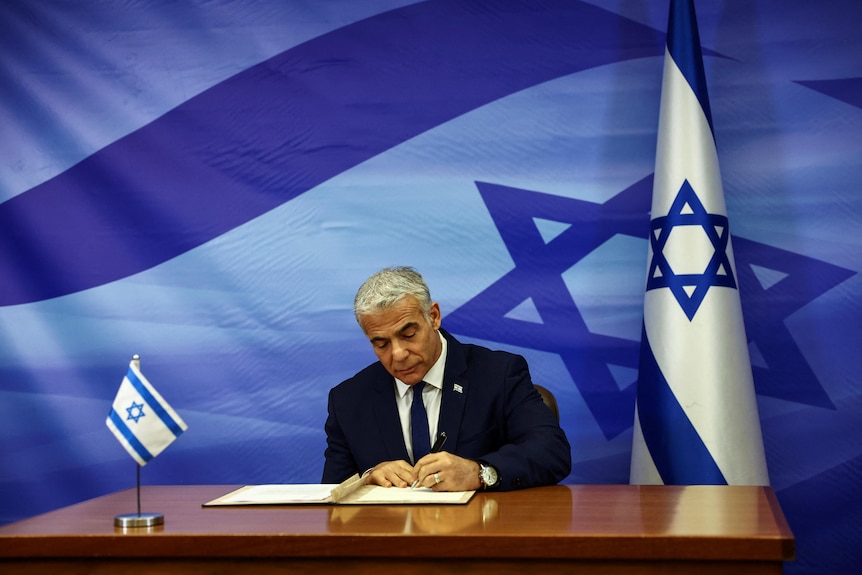 Ronen Zvulun signs on paper sitting on a desk, in front of the national flag imagery.
