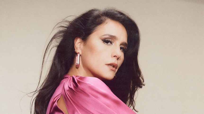 Jessie Ware looks over her shoulder, hair flowing, wearing a pink silk dress against a light brown backdrop