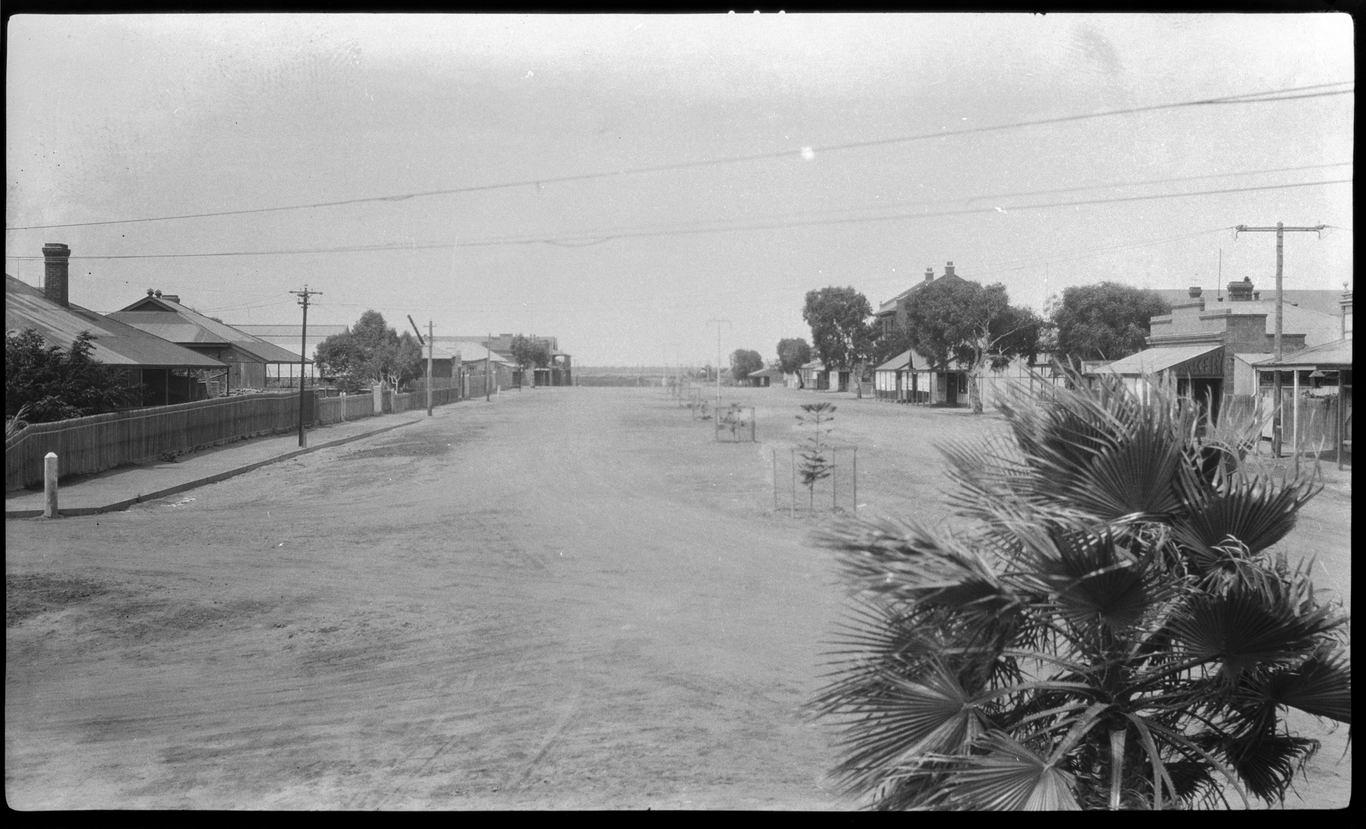 A black and white photo of a wide dirt road lined with houses, with a palm tree in the corner right foreground.