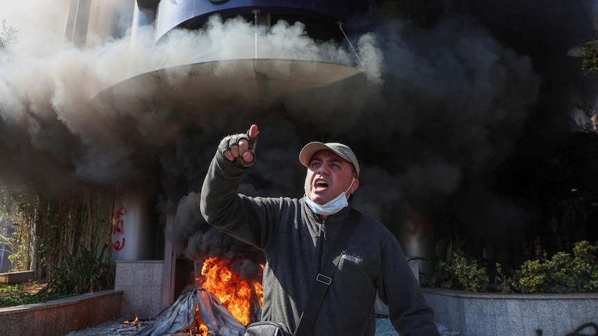 A man with a arm raised in front of a smoldering building with visible flames