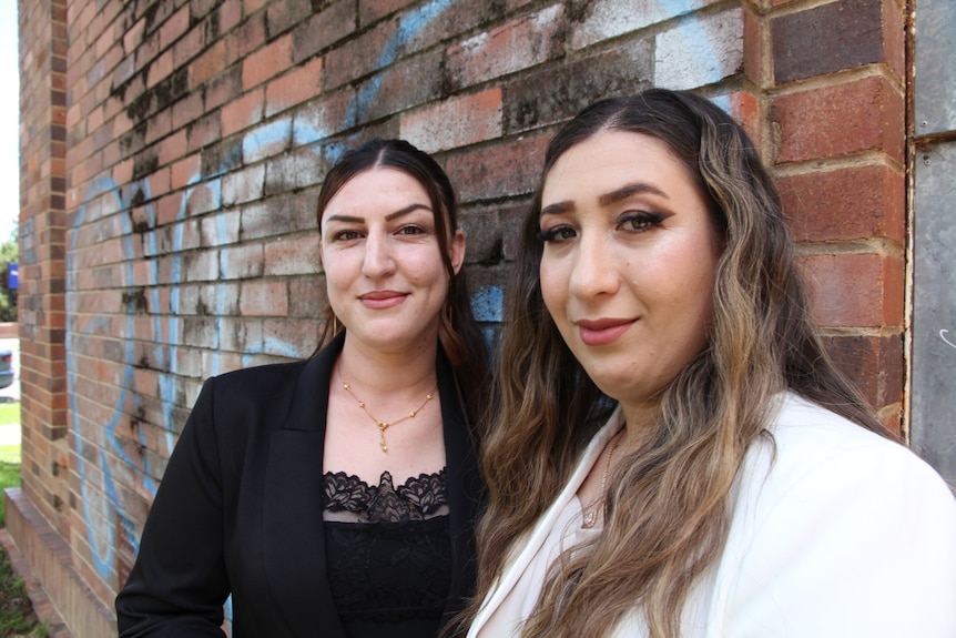 Two well-dressed young women of Middle Eastern appearance lean against a brick wall and smile at the camera.   