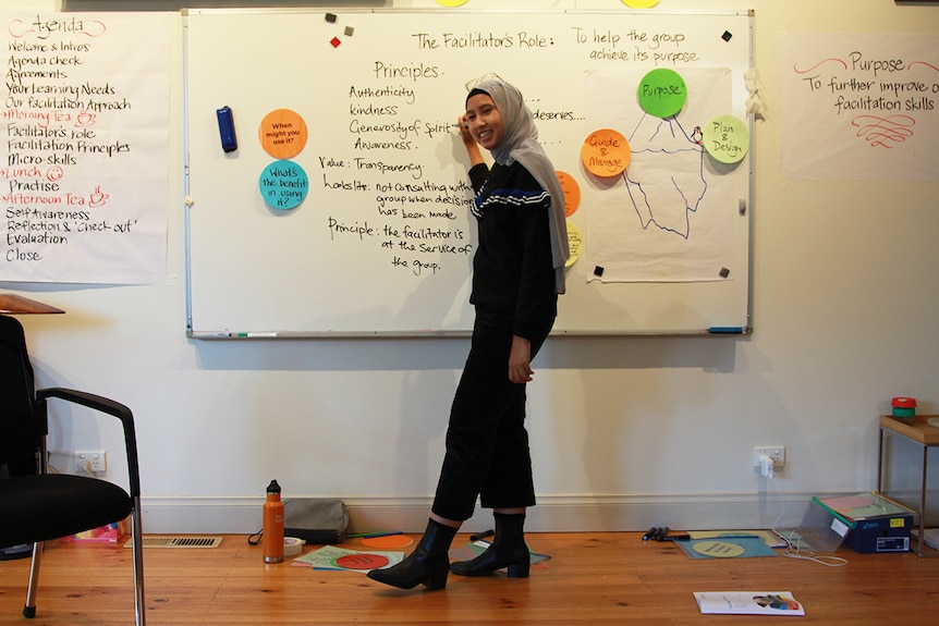 A young woman wearing a light grey hijab and a black jumper stands in front of a whiteboard.