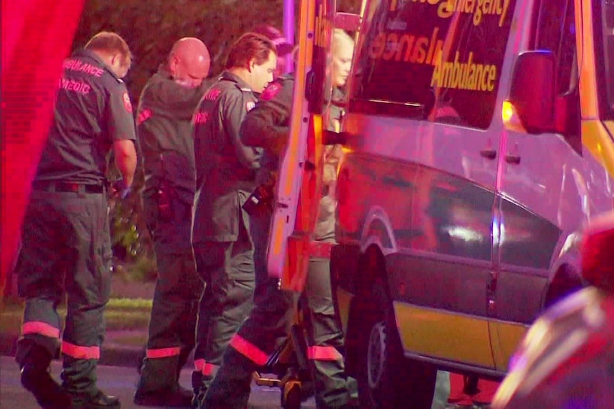 Paramedics in green uniforms surrounding the back of an ambulance with lights flashing
