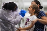 A girl sits on a chair with a woman behind her as a person dressed in scrubs puts a swab test in the girls mouth